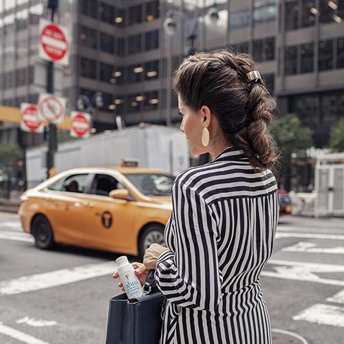 Women walking through a New York road with orange taxi on her right and keeping the rahua voluminous dry shampoo bottle inside her black purse