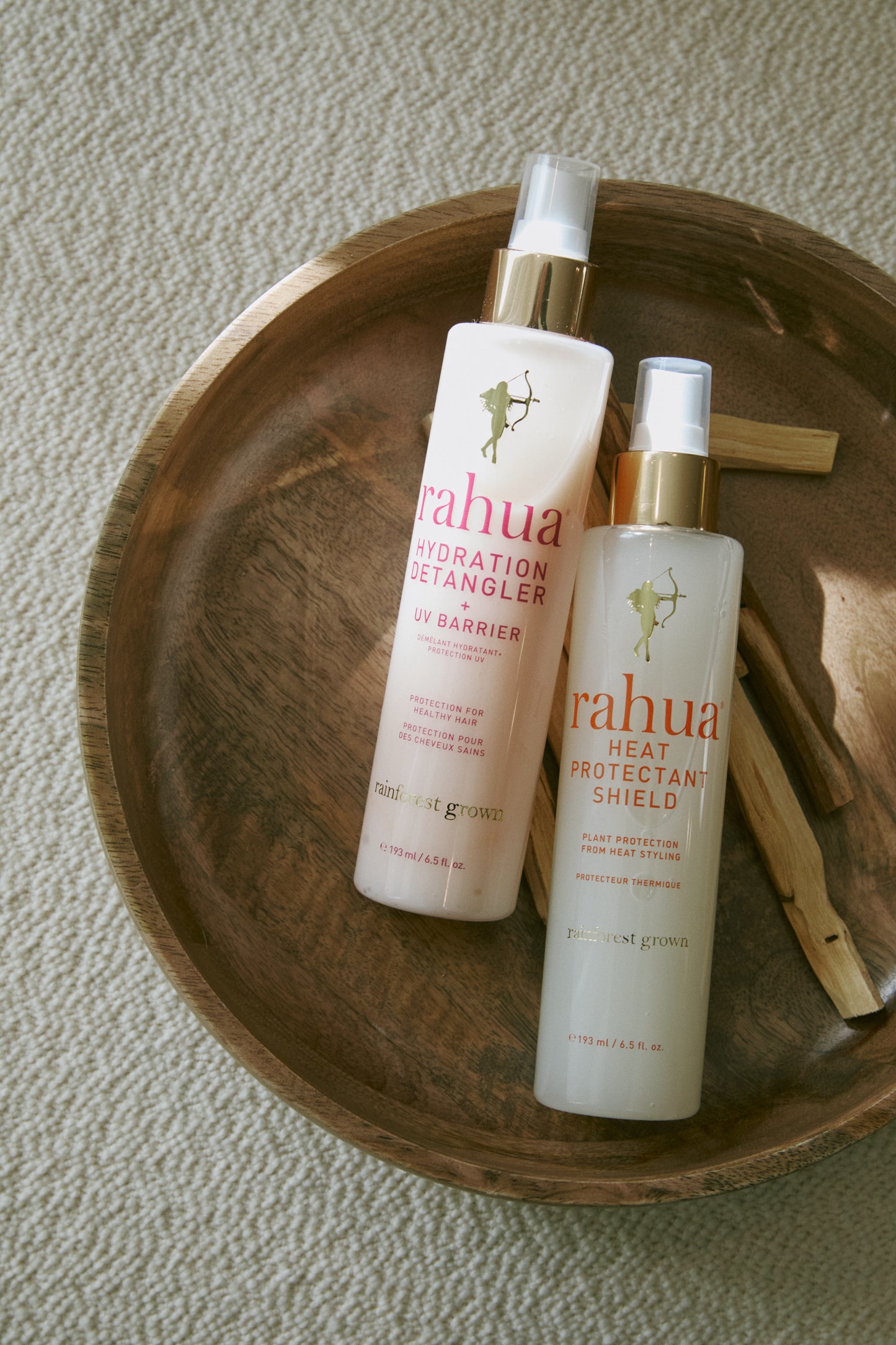 two bottles of a rahua hydration detangler and heat protectant shield in a wooden bowl with sticks of palo santo incense on a off white fabric background|variant:2-oz