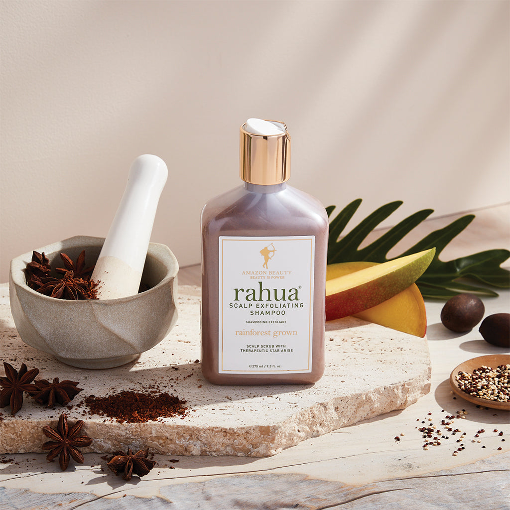 bottle of rahua scalp exfoliating shampoo sitting on a stone slab with a plain beige background with mangoes, quinoa, and star anise in a mortar and pestle being crushed|variant:074-oz