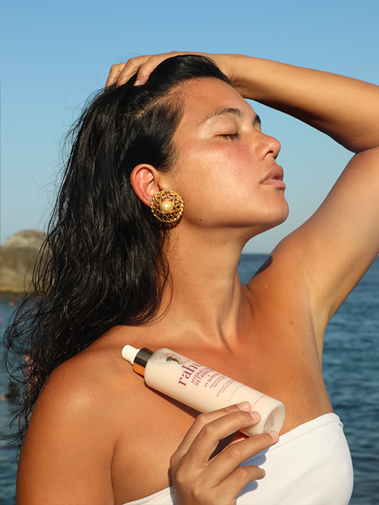 model posing in the sun by the seaside with a bottle of rahua hydration detangler and uv barrier|variant:2-oz