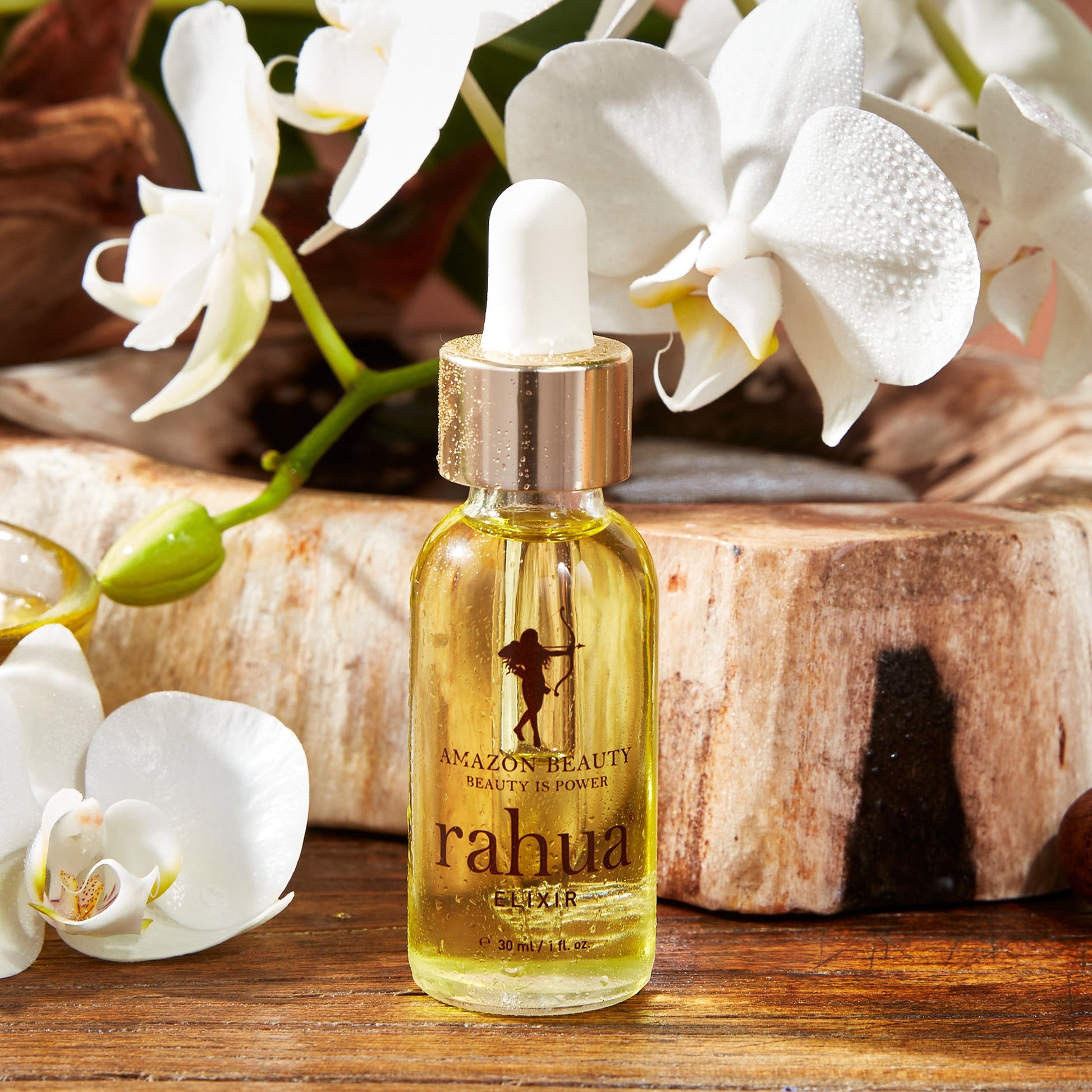 bottle of golden liquid in a glass bottle rahua elixir oil made of 100% rahua oil on a plain white background on a wooden table with orchids in the background|variant:1-oz